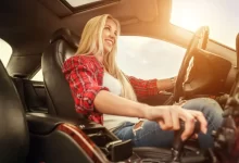 Řidička jede za slunného dne, levou rukou drží volant, pravou rukou ovládá automatickou převodovku