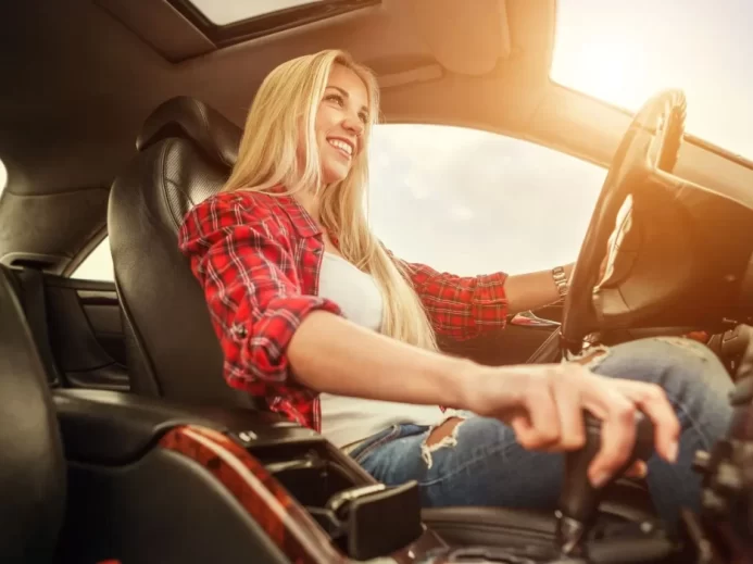 Řidička jede za slunného dne, levou rukou drží volant, pravou rukou ovládá automatickou převodovku