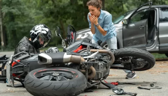 Motorkář po nehodě sedí u své motorky, zatímco žena reaguje na situaci