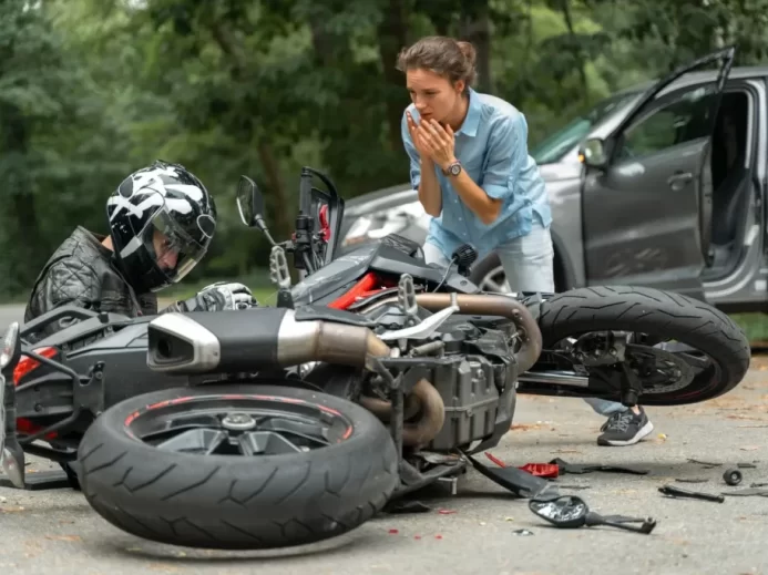 Motorkář po nehodě sedí u své motorky, zatímco žena reaguje na situaci