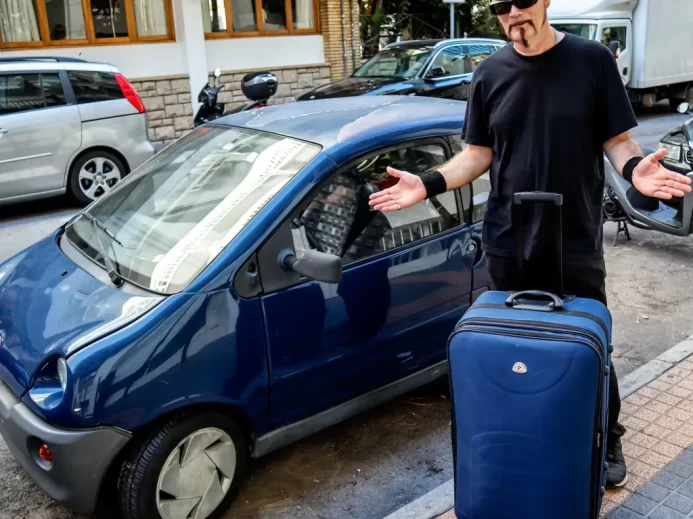 Velký muž v černém s kufrem u malého modrého auta