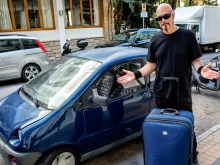 Velký muž v černém s kufrem u malého modrého auta