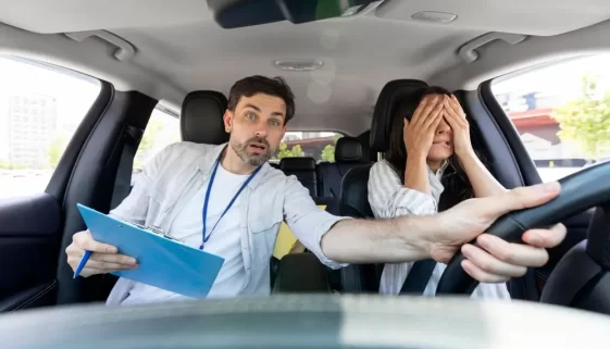 Žena si při řízení auta zakrývá oči, instruktor autoškoly drží volant