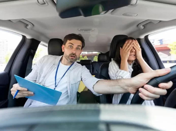 Žena si při řízení auta zakrývá oči, instruktor autoškoly drží volant