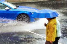Dva jdou lidé pod modrým deštníkem, v pozadí po mokré vozovce přejíždí modré auto.