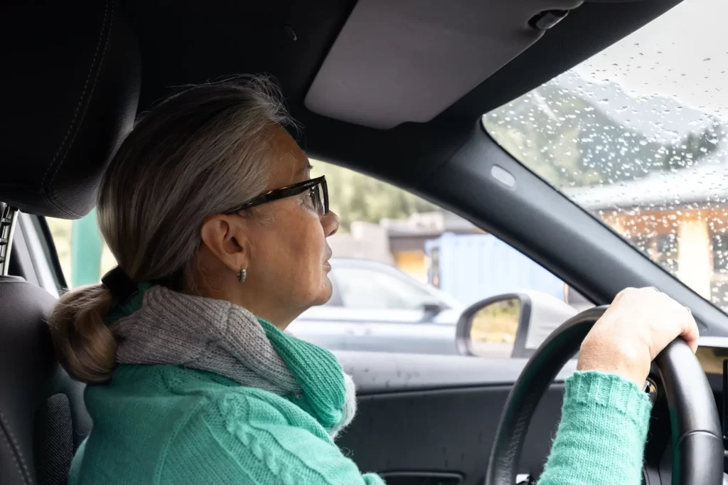 - MotorGuru.cz STarší žena v zeleném oblečení řídí auto v dešti - detail z kabiny vozu.