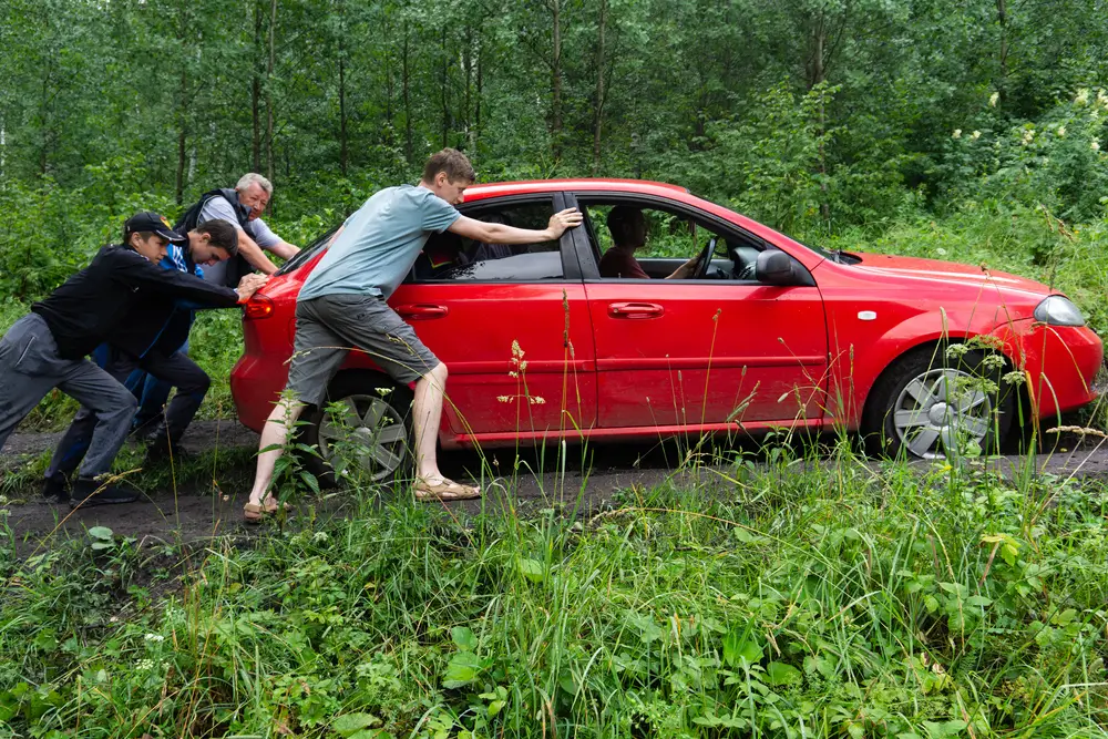 Skupina lidí tlačí červené osobní auto uvízlé v bahně na lesní cestě 