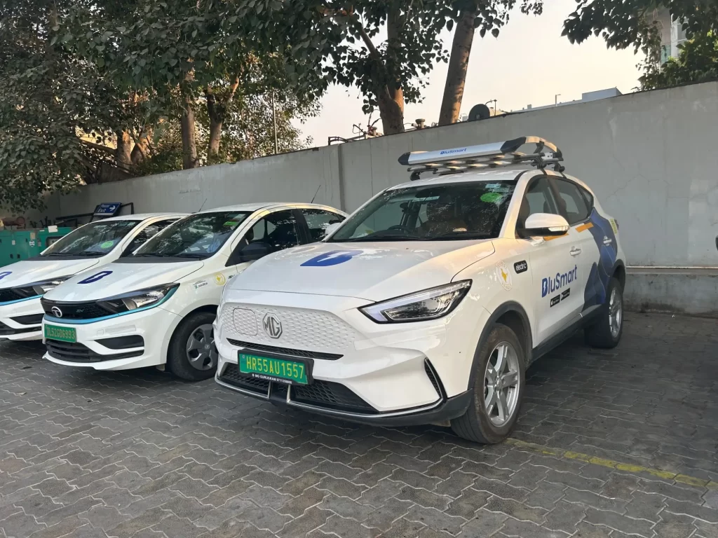 Několik zaparkovaných elektrických vozů společnosti BluSmart, včetně SUV značky MG a sedanů Tata. Všechny vozy mají bílé karoserie s modrým logem BluSmart a zelenými registračními značkami.