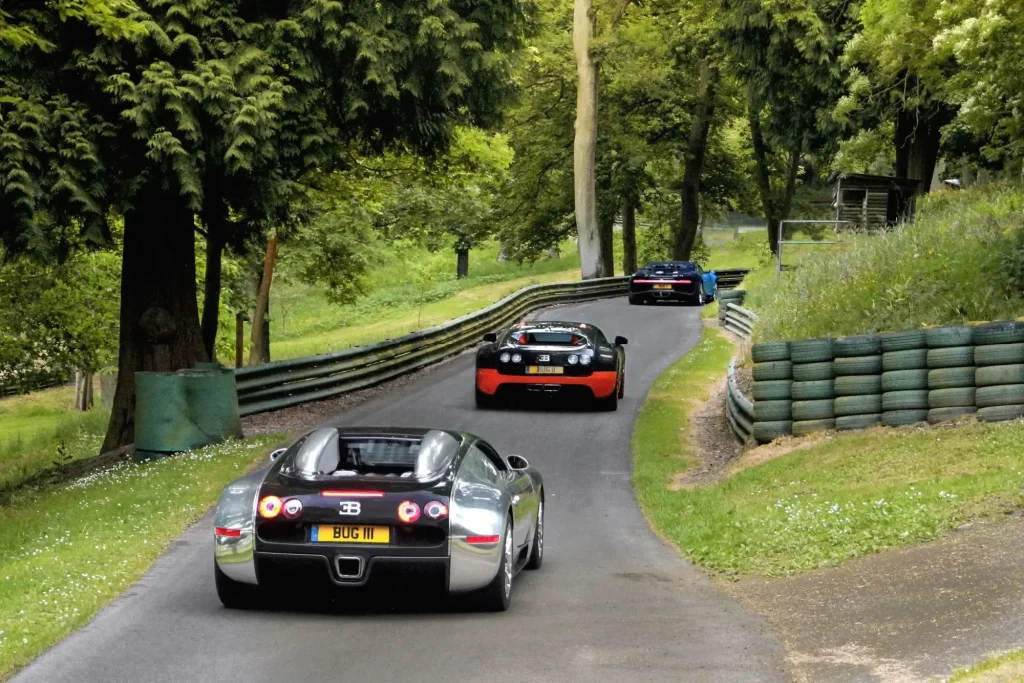 Tři vozy značky Bugatti při jízdě po úzké silnici obklopené přírodou.