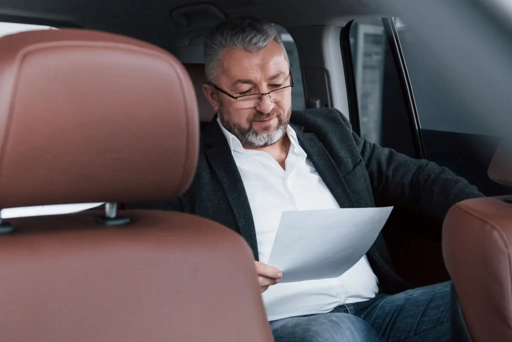 Muž ve středním věku sedí na zadním sedadle automobilu, oblečený v elegantním saku a bílé košili, a soustředěně čte dokument.