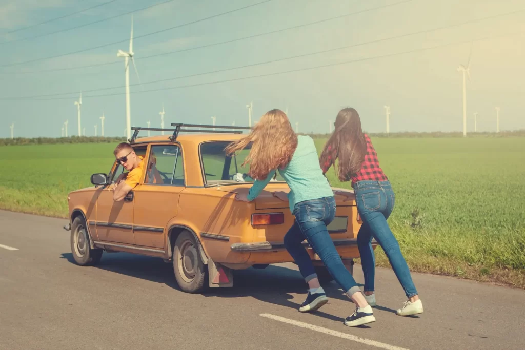 Dvě mladé ženy tlačí starší žluté auto, muž sedí na místě řidiče. 