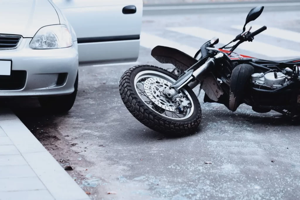 Dopravní nehoda mezi automobilem a motocyklem. Přední část motorky leží převrácená na zemi přímo vedle otevřených dveří zaparkovaného vozu.