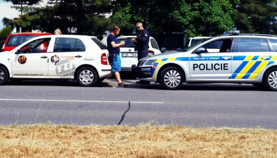 Policista právě kontroluje doklady řidiče bílého služebního vozu s logem TD servis, zatímco policejní vozidlo stojí zaparkované hned vedle.
