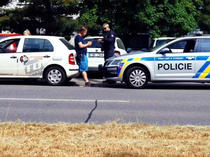 Policista právě kontroluje doklady řidiče bílého služebního vozu s logem TD servis, zatímco policejní vozidlo stojí zaparkované hned vedle.