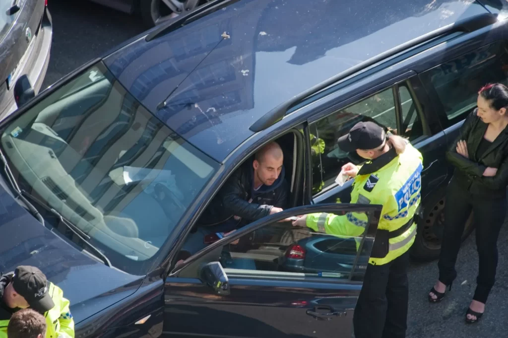 Policista v reflexní vestě kontroluje doklady řidiče tmavého osobního vozu, který zůstává sedět ve voze a podává doklady otevřeným okénkem.