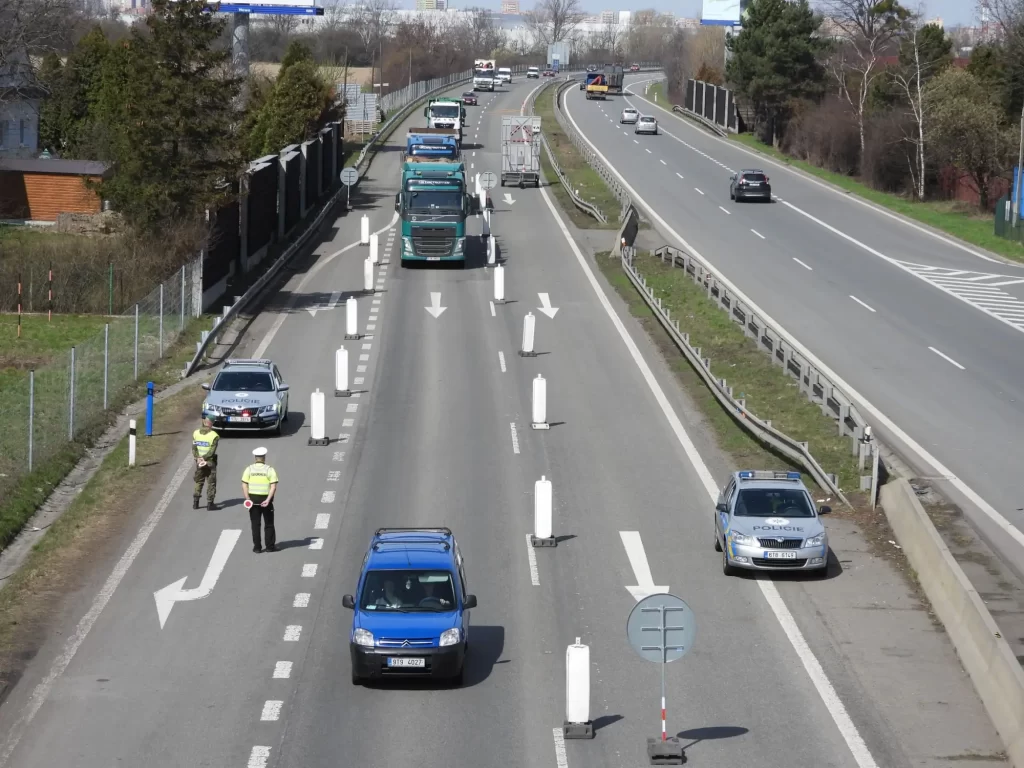 Na silnici jsou rozmístěny plastové směrovací kužely, které vytvářejí koridor pro kontrolu vozidel. Na kraji silnice stojí policejní vozy a dva policisté, kteří sledují projíždějící auta. 