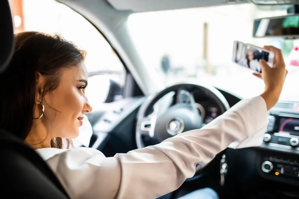 Mladá žena sedí na místě řidiče v moderním autě a fotí si selfie telefonem. 