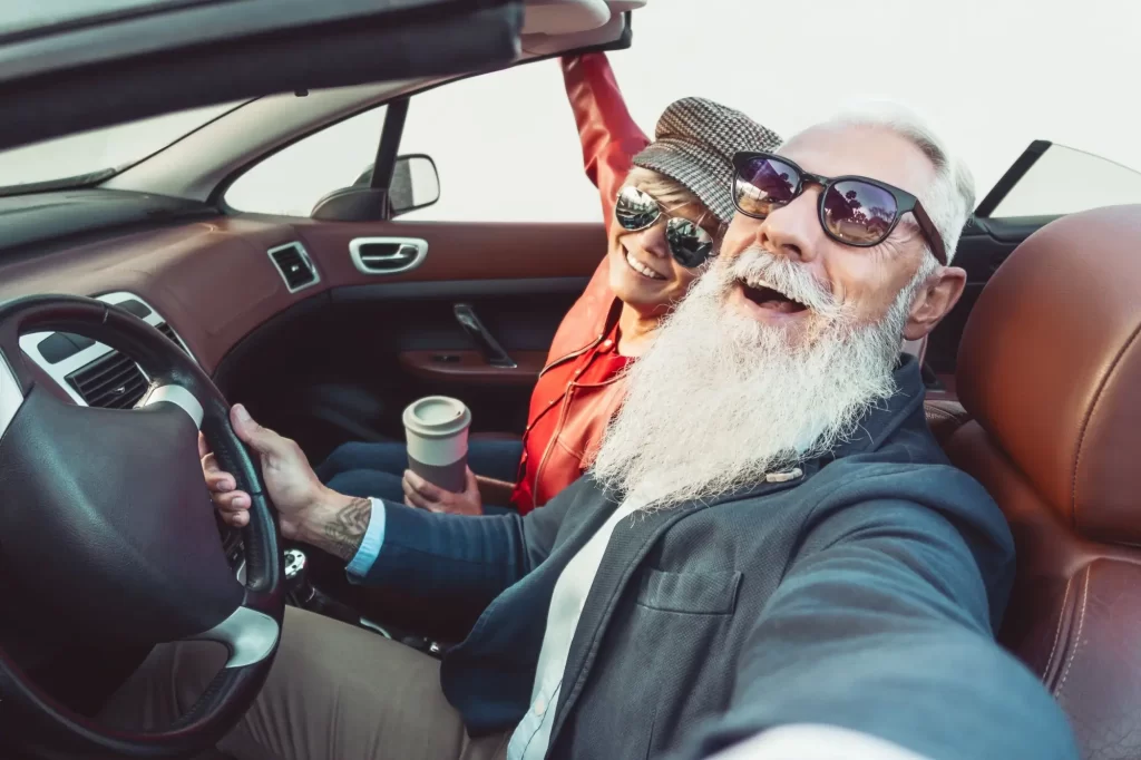 Muž s dlouhým bílým plnovousem a tmavými brýlemi drží volant i telefon a pořizuje selfie. 