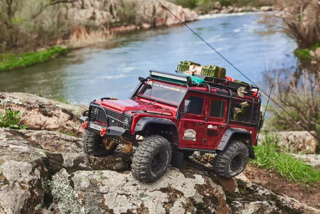 RC model vozu Land Rover Defender v červené barvě, který zdolává skalnatý terén u řeky