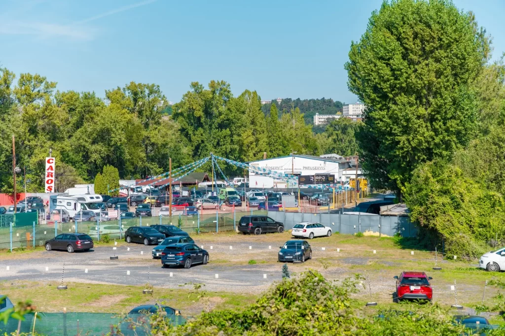 - MotorGuru.cz Shluk zaparkovaných vozidel – především ojetých osobních aut – obklopený plotem a barevnými praporky.