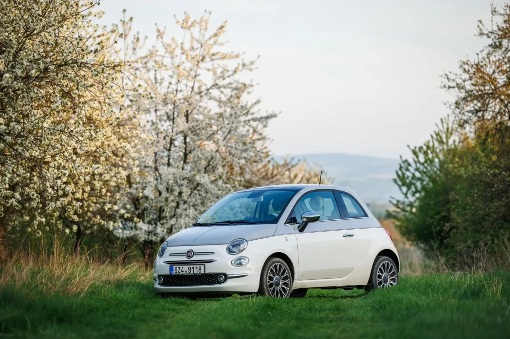 - MotorGuru.cz Fiat 500, zaparkovaný na zelené louce uprostřed kvetoucích stromů.