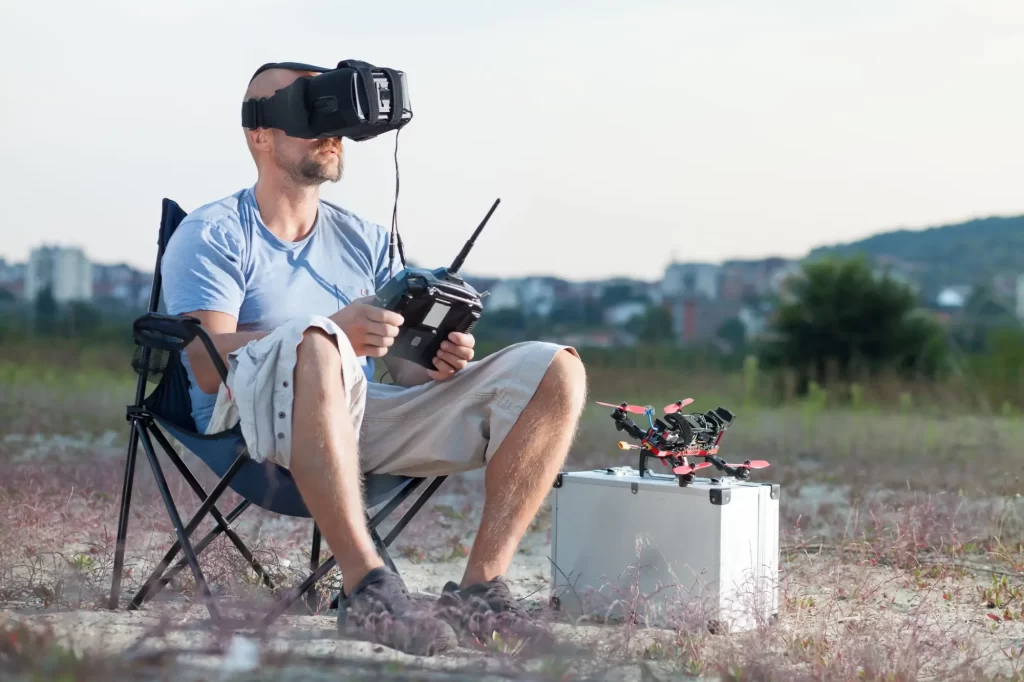 - MotorGuru.cz Pilot FPV dronu během tréninku v přírodě. Sedí na skládací židličce, má nasazené FPV brýle a v rukou drží dálkový ovladač s anténami.