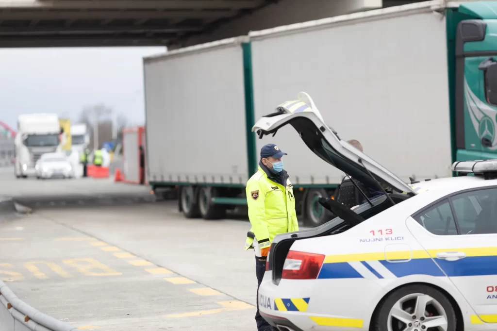 V popředí stojí policejní vůz s otevřeným kufrem, u kterého hlídkuje policista v reflexní bundě a roušce