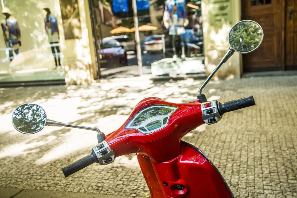 - MotorGuru.cz Červený skútr značky Vespa, zaparkovaný na městské dlažbě před výlohou butiku.