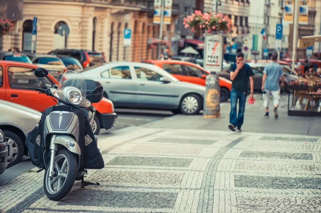 - MotorGuru.cz Stříbrný skútr zaparkovaný na typické pražské mozaikové dlažbě, těsně u přechodu a rušné ulice plné aut.