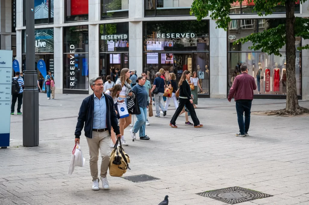 Lidé různých věkových kategorií procházejí kolem obchodů, nesou nákupní tašky nebo si jen užívají klidného prostoru bez automobilového provozu. 