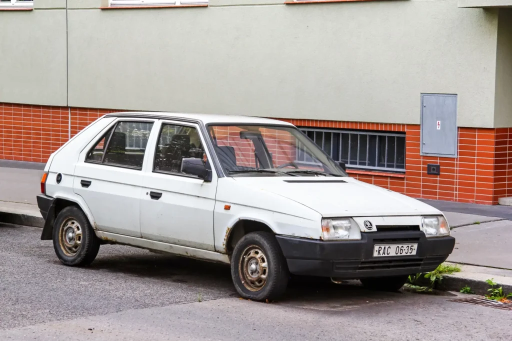 Bílá Škoda Favorit zaparkovaná u chodníku, s viditelnými známkami koroze na prazích a kolech.