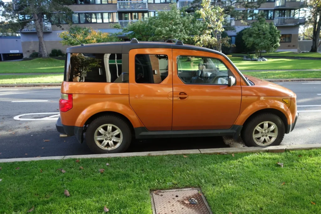 Oranžová Honda Element zaparkovaná u obrubníku v městské ulici.