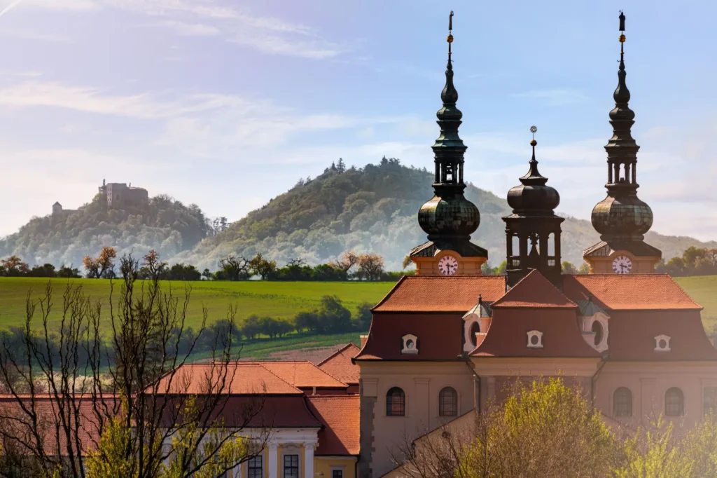 Poutní místo s barokním kostelem se třemi věžemi, v pozadí se zvedají zalesněné kopce s hradem.