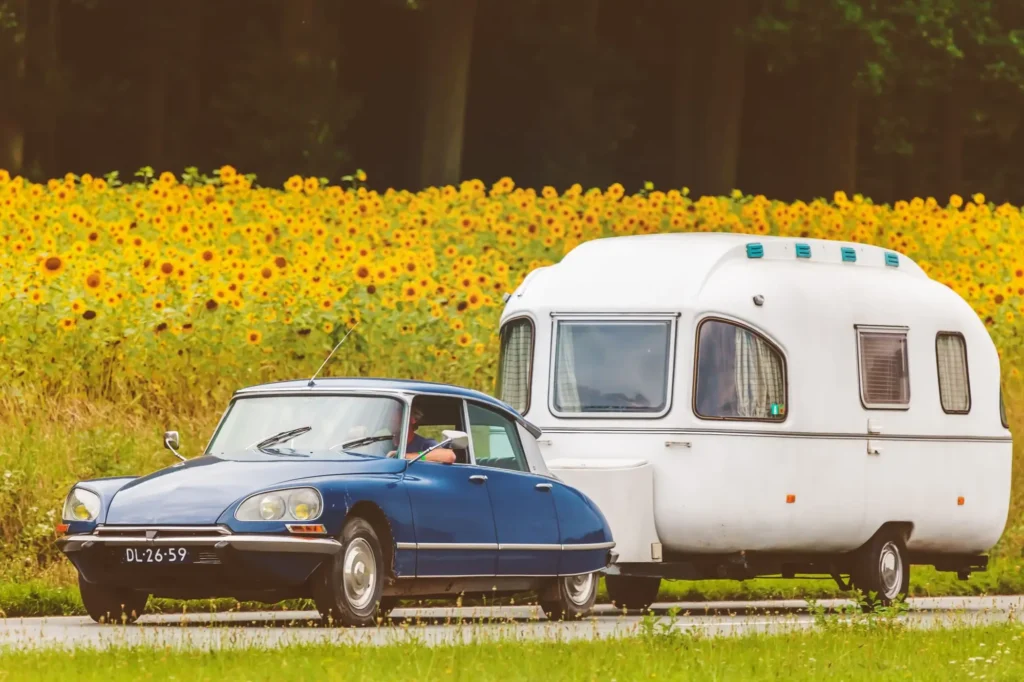 - MotorGuru.cz Klasický automobil Citroën DS, který táhne bílý karavan po silnici lemované rozkvetlým polem slunečnic.