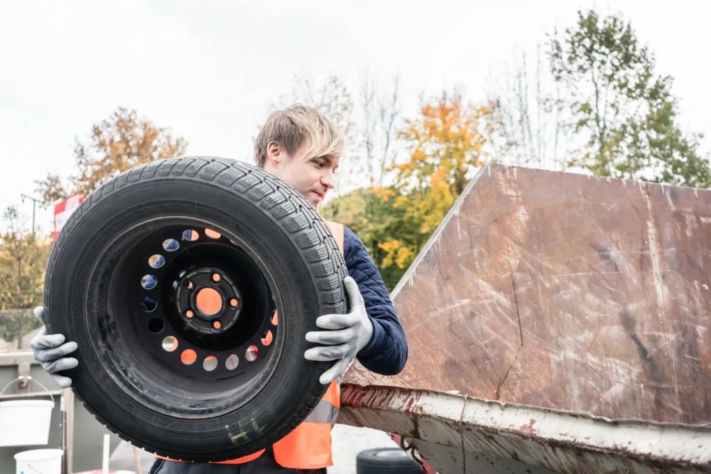 - MotorGuru.cz Muž v reflexní vestě drží starou pneumatiku a vkládá ji do velkého kovového kontejneru.