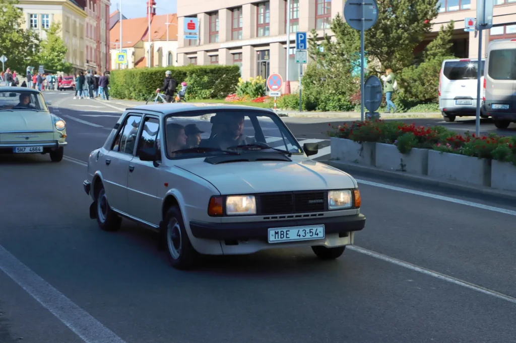 - MotorGuru.cz Bílá Škoda 120 jedoucí městskou ulicí během srazu veteránů, za ní je vidět další historické auto a několik lidí sledujících akci.