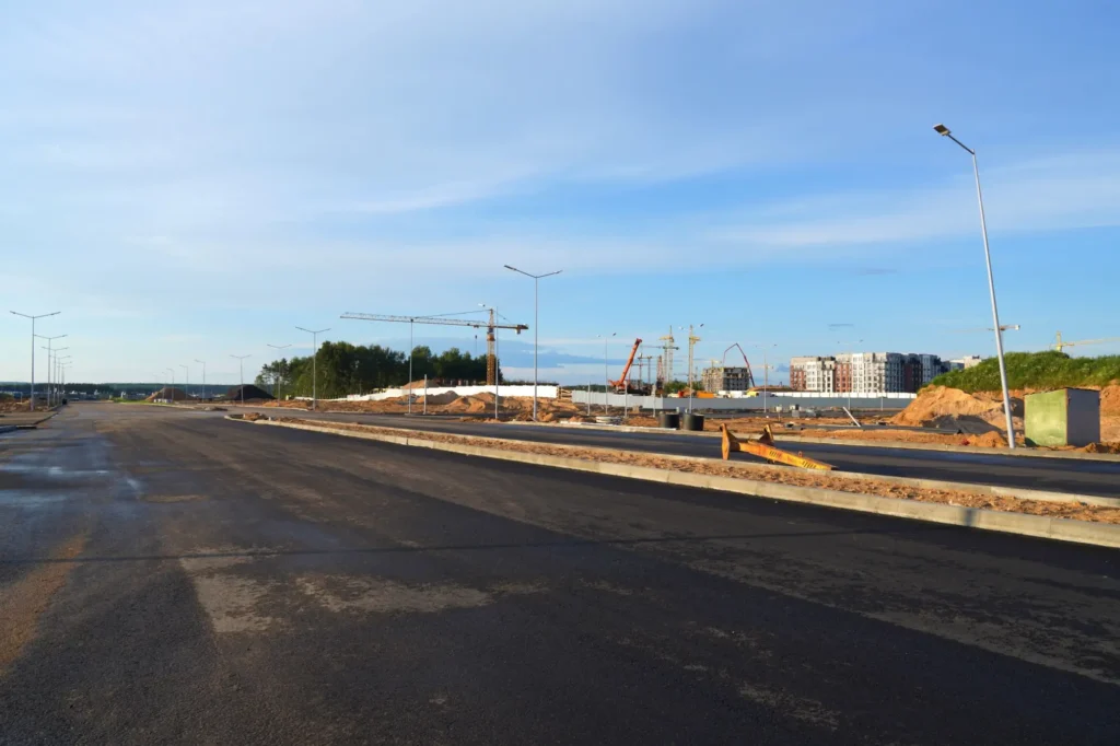 Rozestavěná silnice se zbrusu novým asfaltem, pouličními lampami a v pozadí se staví domy i další infrastruktura.
