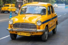Ikonické žluté taxi typu Hindustan Ambassador, jedoucí po městské silnici v Indii.