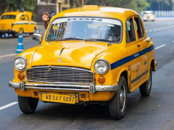 Ikonické žluté taxi typu Hindustan Ambassador, jedoucí po městské silnici v Indii.